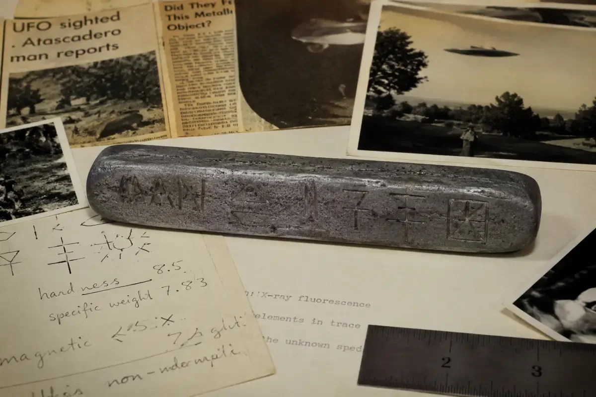 AI illustration of a metal bar engraved with the word "MAN" and unknown symbols, resting on a research desk surrounded by documents and newspaper clippings from the alleged 1970s UFO case.